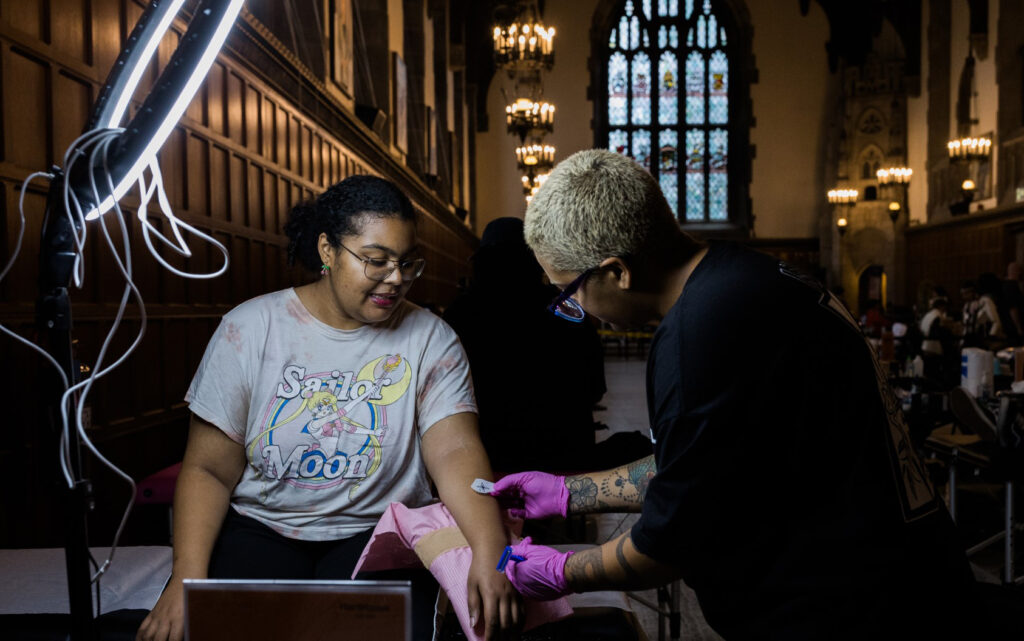 A female student getting a tattoo from a tattoo artist in the Great Hall.