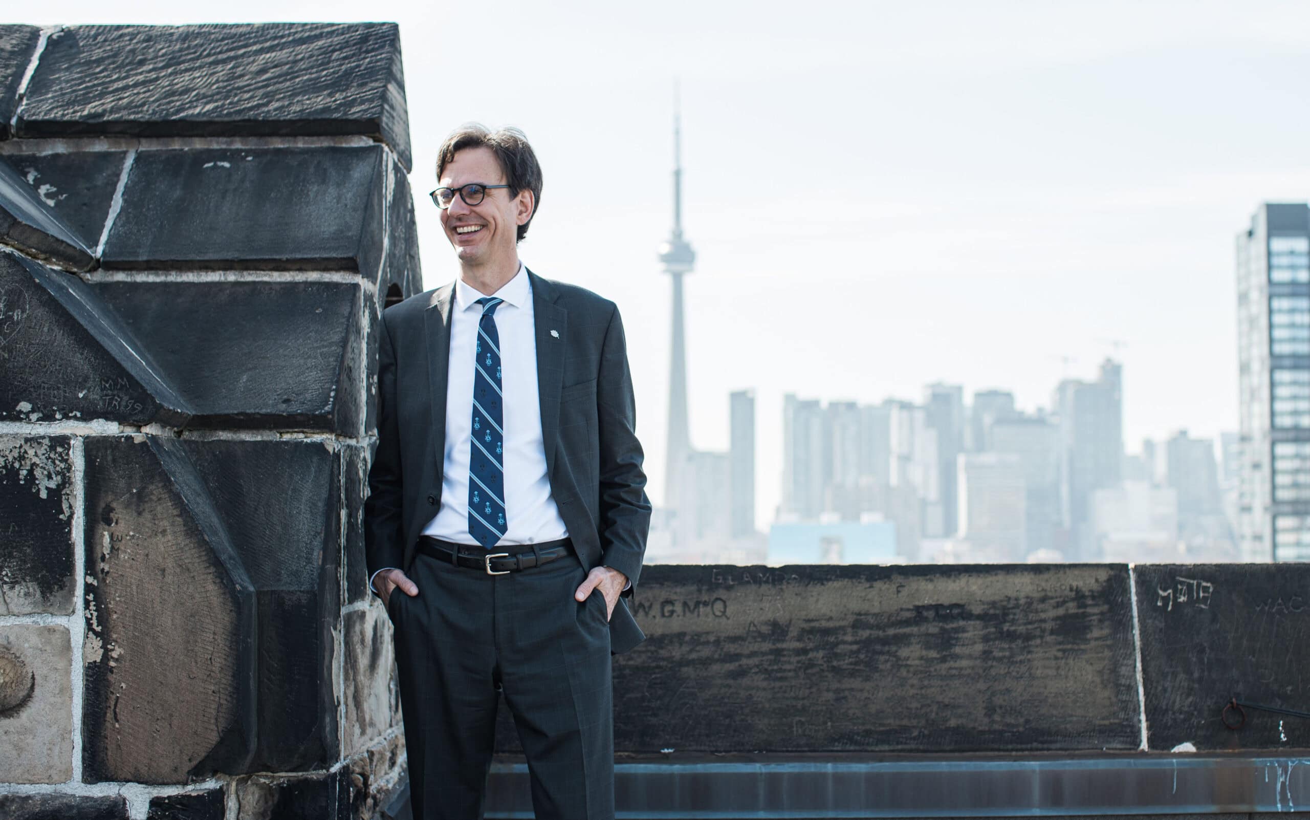 Corwin Cambray stands on a rooftop patio wearing a suit, smiling, with the CN Tower in the background.