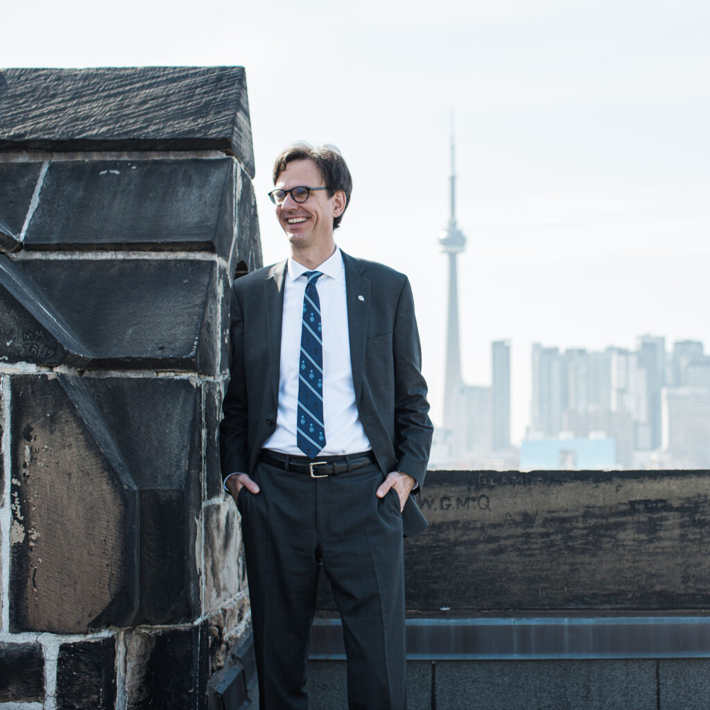 Corwin Cambray wearing a suit, smiling, standing on a rooftop balcony with the CN Tower in the background.