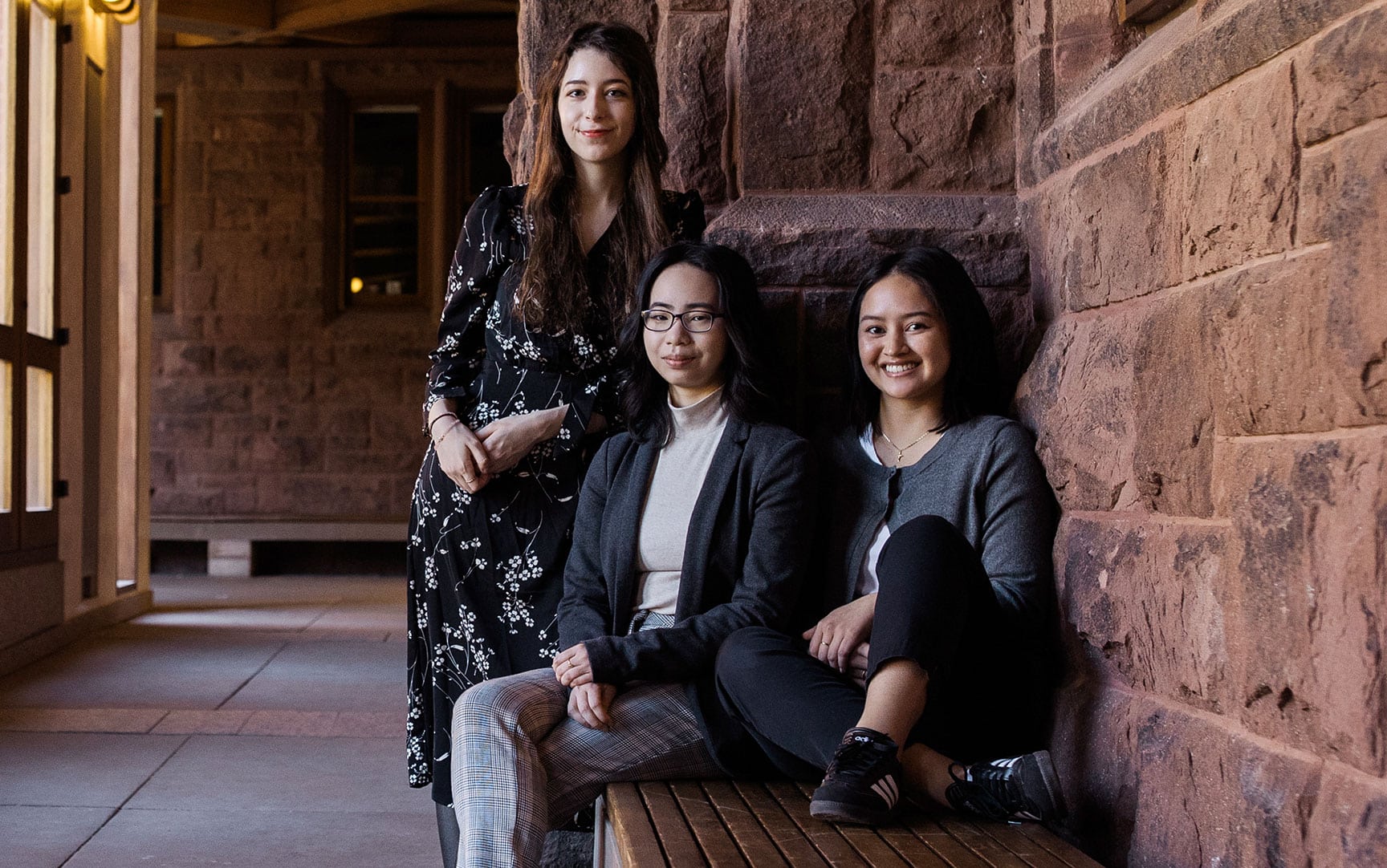 Three female students sit together next to a brick wall at the Asian Institute.