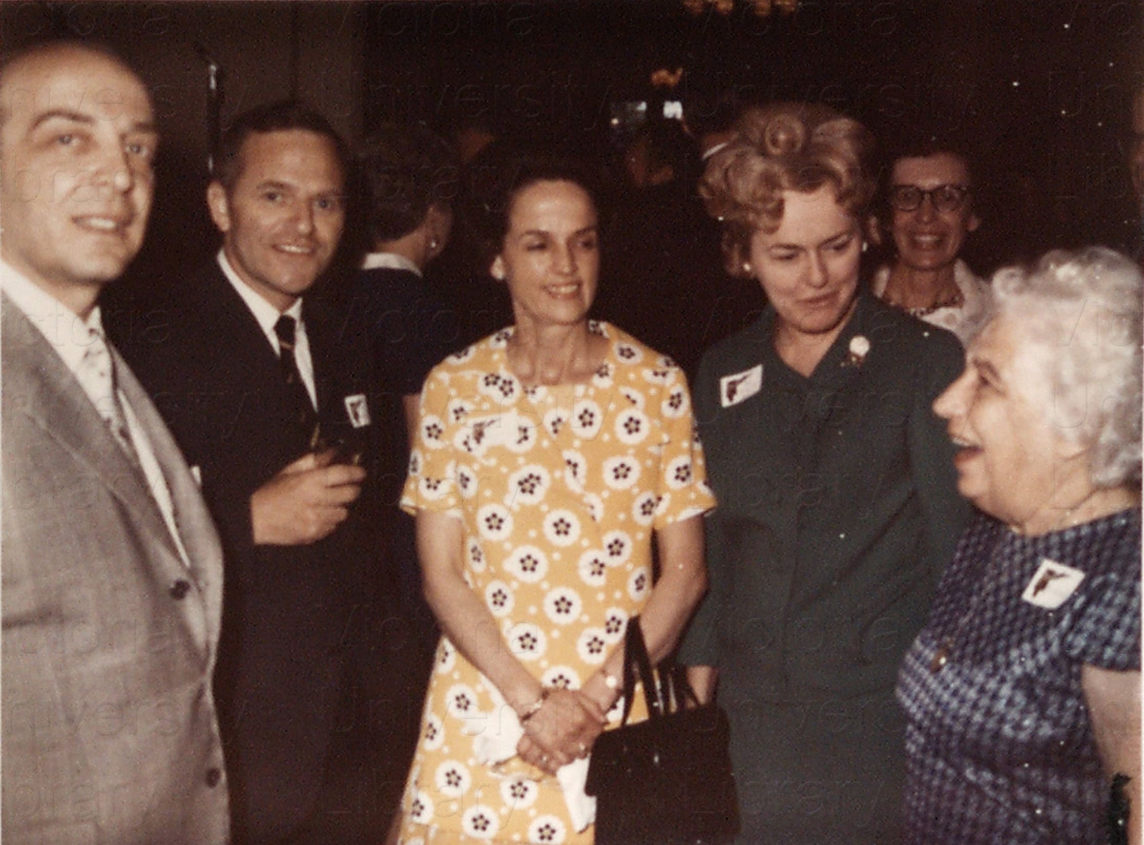 five people gathered at a party in formal wear, smiling at the camera, as a sixth person looks over