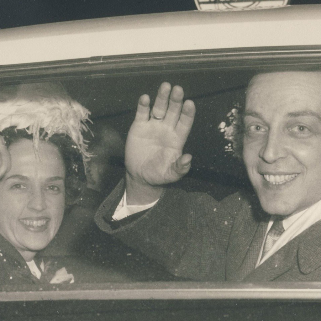 Black and white photo of a couple waving through the back windshield of their car