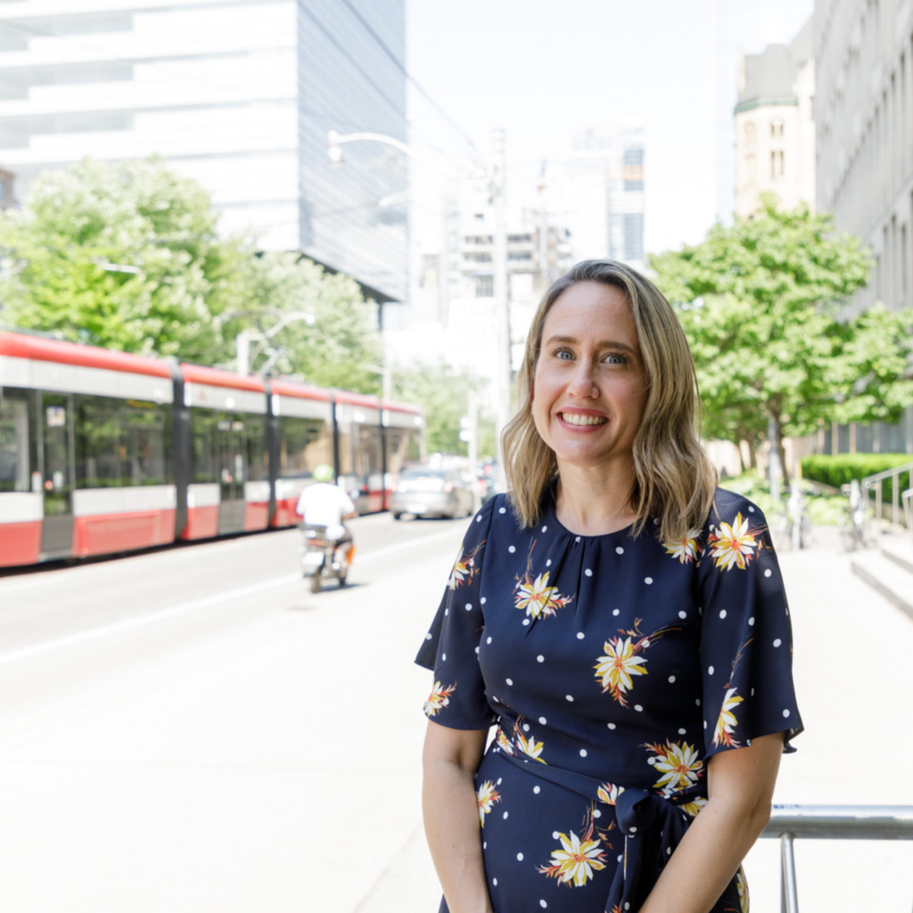 Kristin Cleverly in a blue dress with yellow flowers stands against a bike rack, with a street car in the background