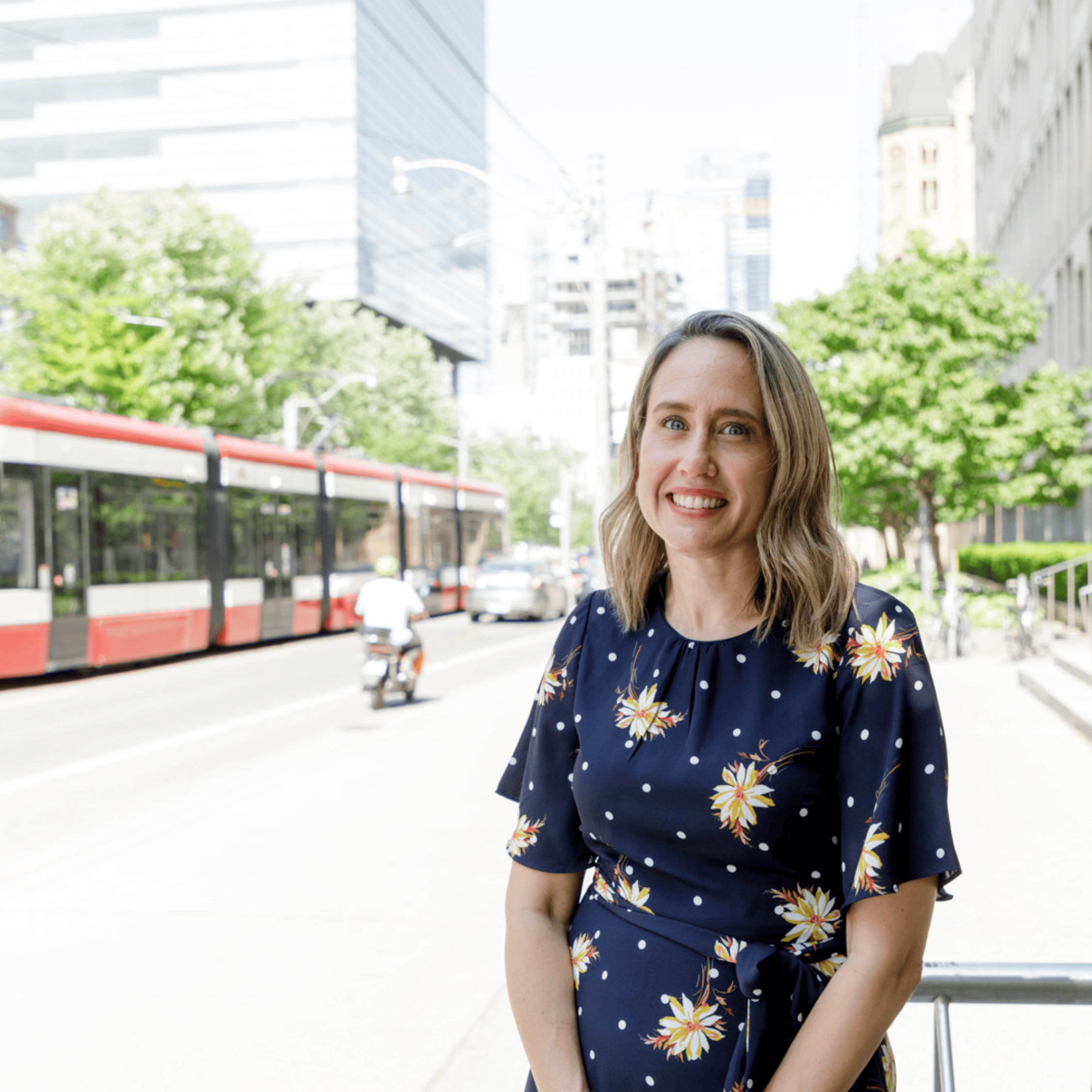 Kristin Cleverly in a blue dress with yellow flowers stands against a bike rack, with a street car in the background