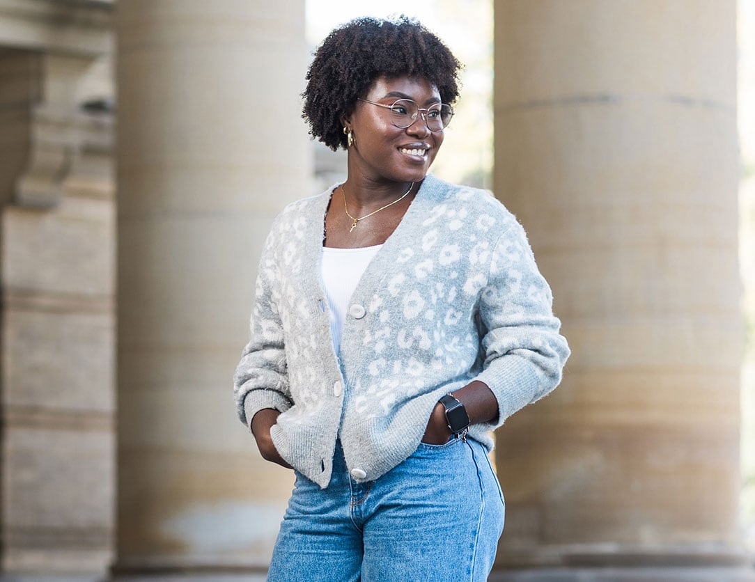 Image of Favour Aina standing in front of a building, wearing glasses, a grey leopard cardigan and blue jeans.