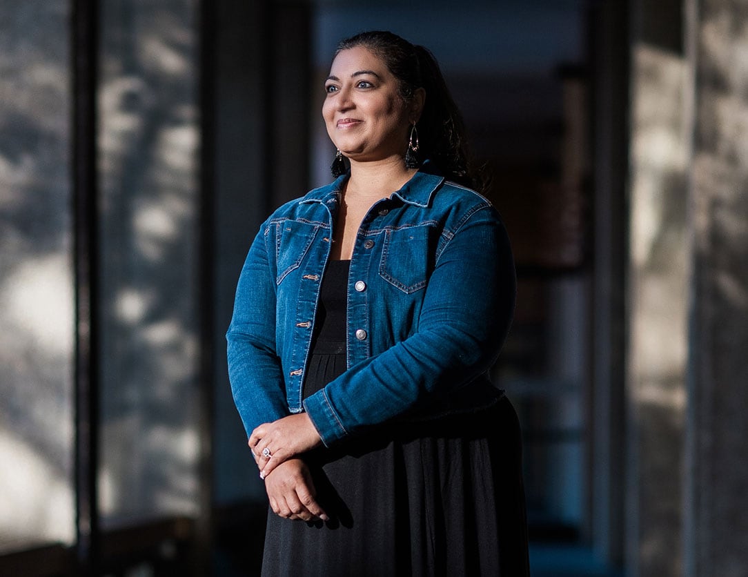 Image of Shezadi Khushal standing in front of a building, wearing a black dress and jean jacket.