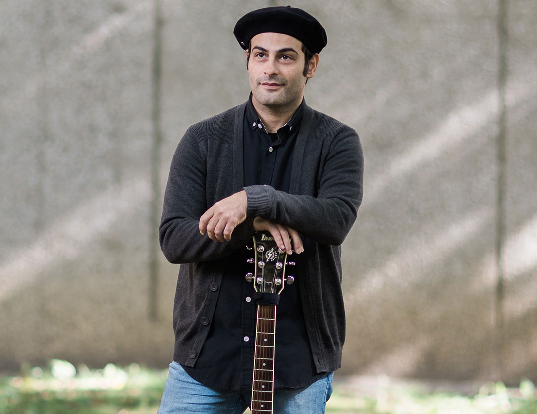 Image of Tony Bali standing outside while leaning on the arm of a guitar, wearing a black beret, black shirt and cardigan.