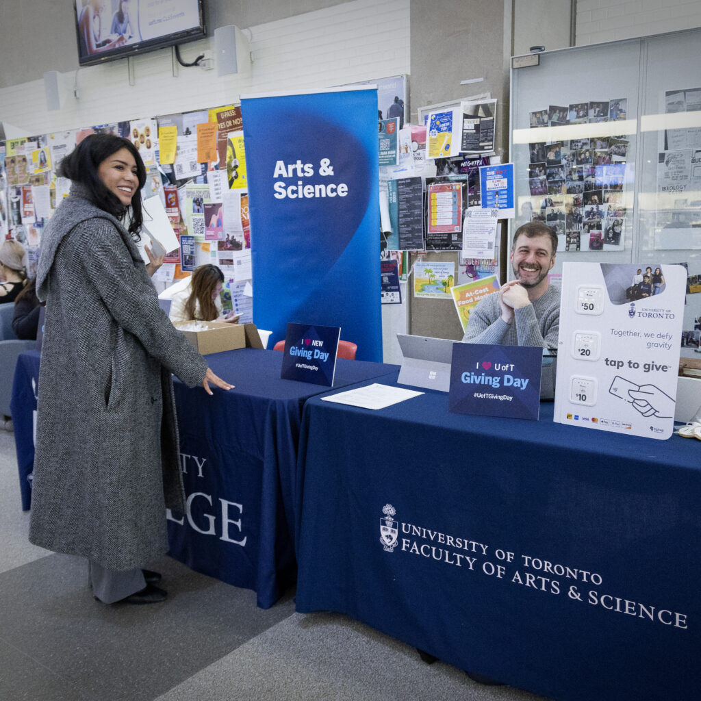 U of T Arts and Science student and staff smiling in Sid Smith Building's lobby, standing next to Giving Day table signage and posters