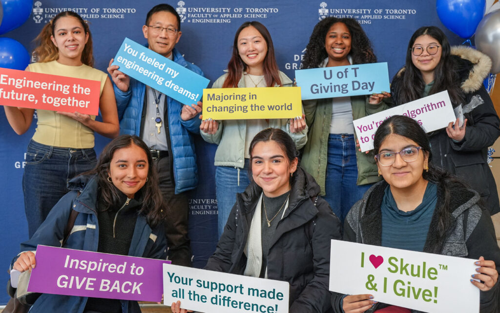 Smiling engineering students posing against a step-and-repeat while holding up signs for U of T Giving Day