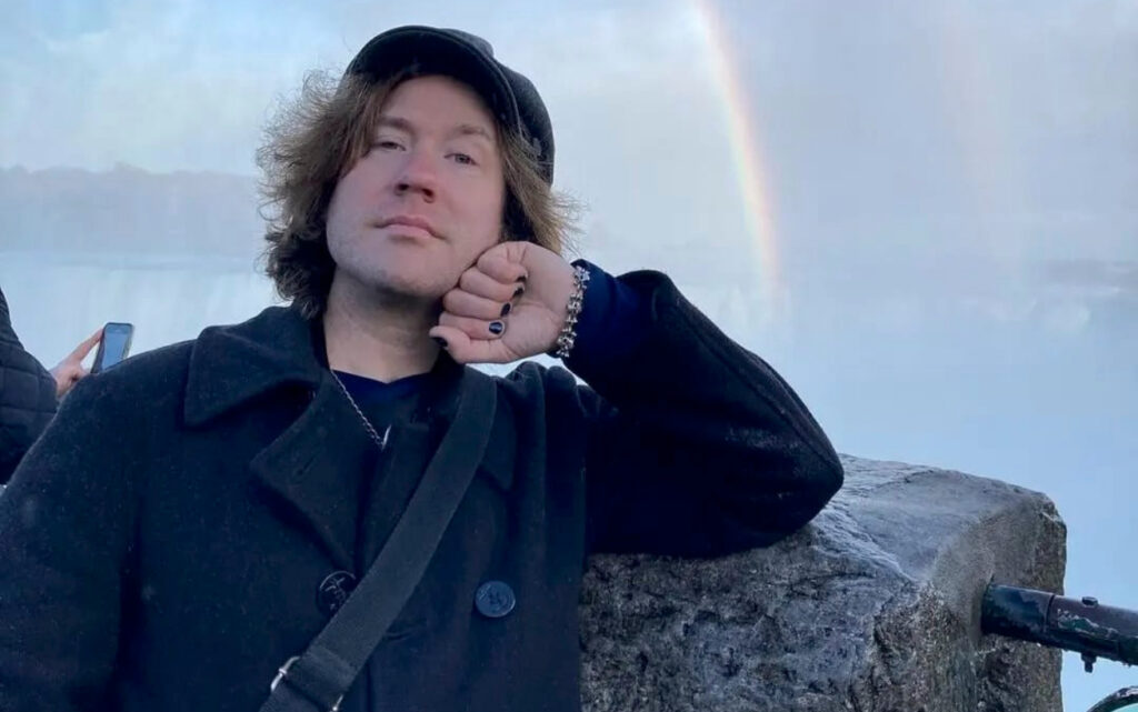 Martin wearing all black. He's posing for a photo in at Niagara Falls and there is a rainbow in the sky behind him.