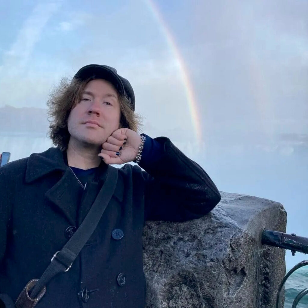 Martin wearing all black. He's posing for a photo in at Niagara Falls and there is a rainbow in the sky behind him.