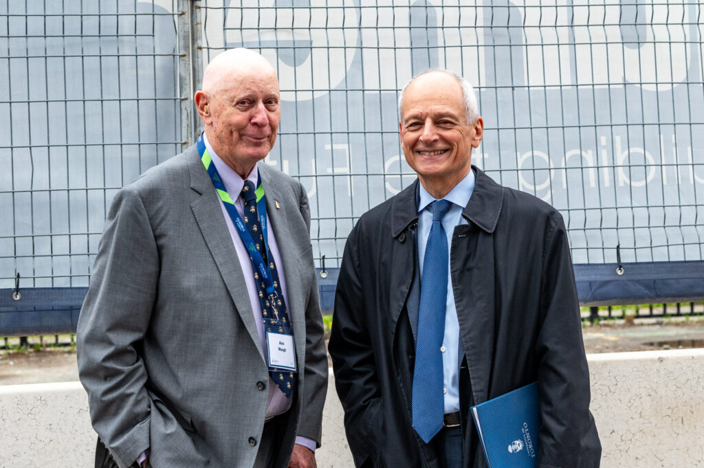 Two men in business suits pose for a photo in front of a construction site. 