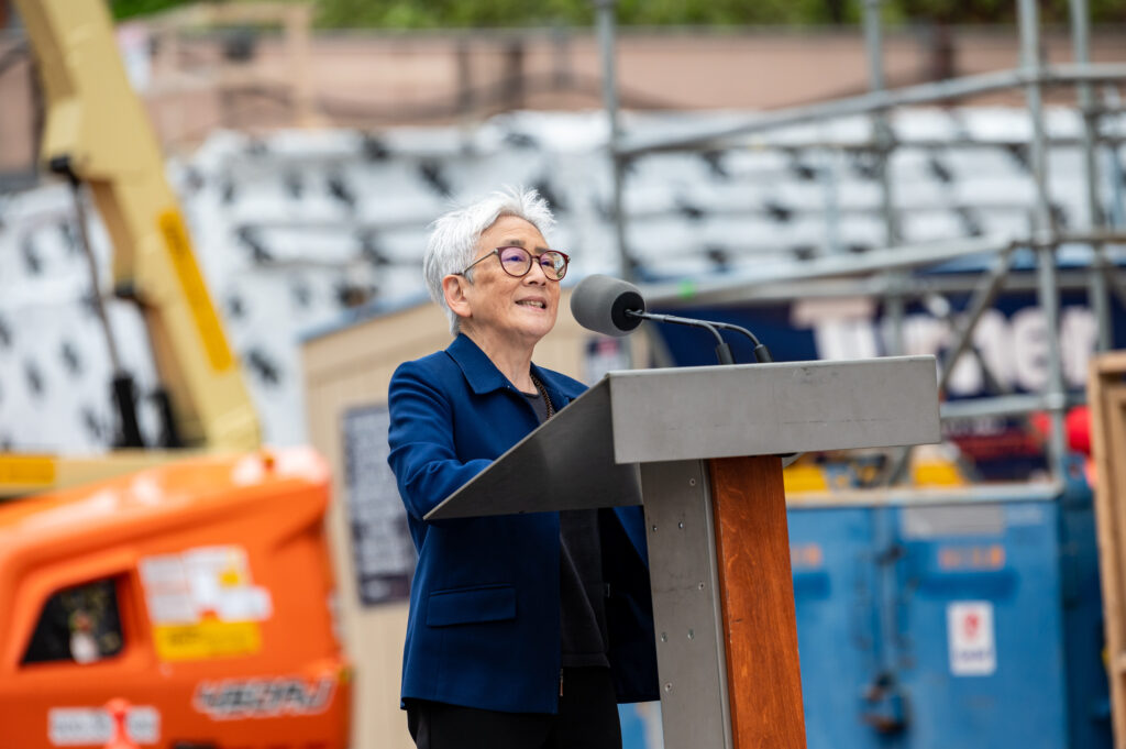 A woman speaks at a podium in front of a construction site. 