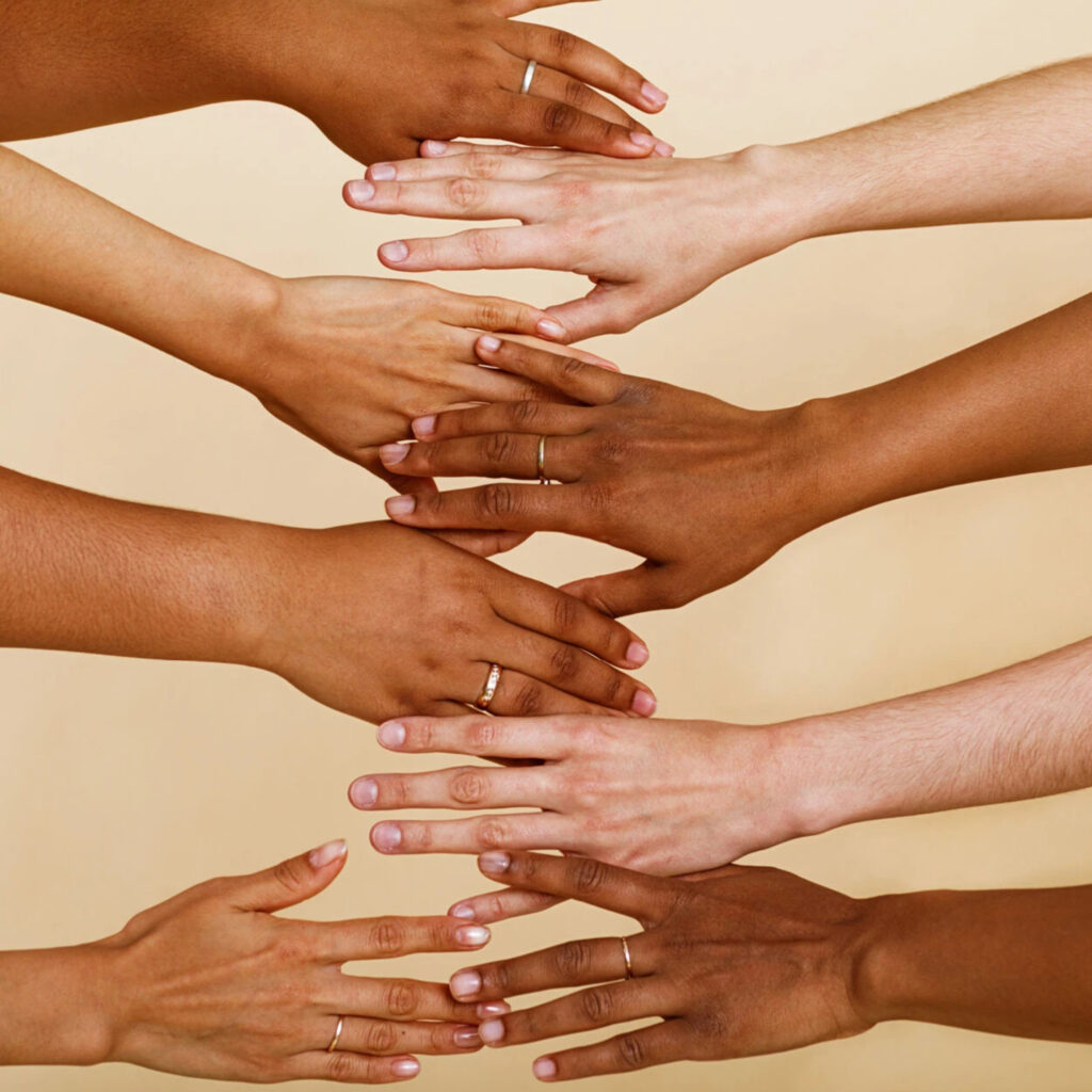 Outstretched hands of multiple different skin tones against a soft yellow backdrop. Some of the hands are wearing simple gold bands.