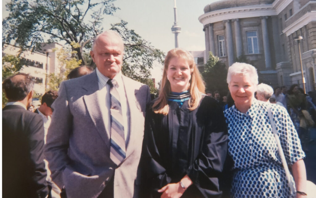 Three people standing in front of Convocation Hall with the CN Tower in the background. Two are parents, one is wearing a graduation gown.