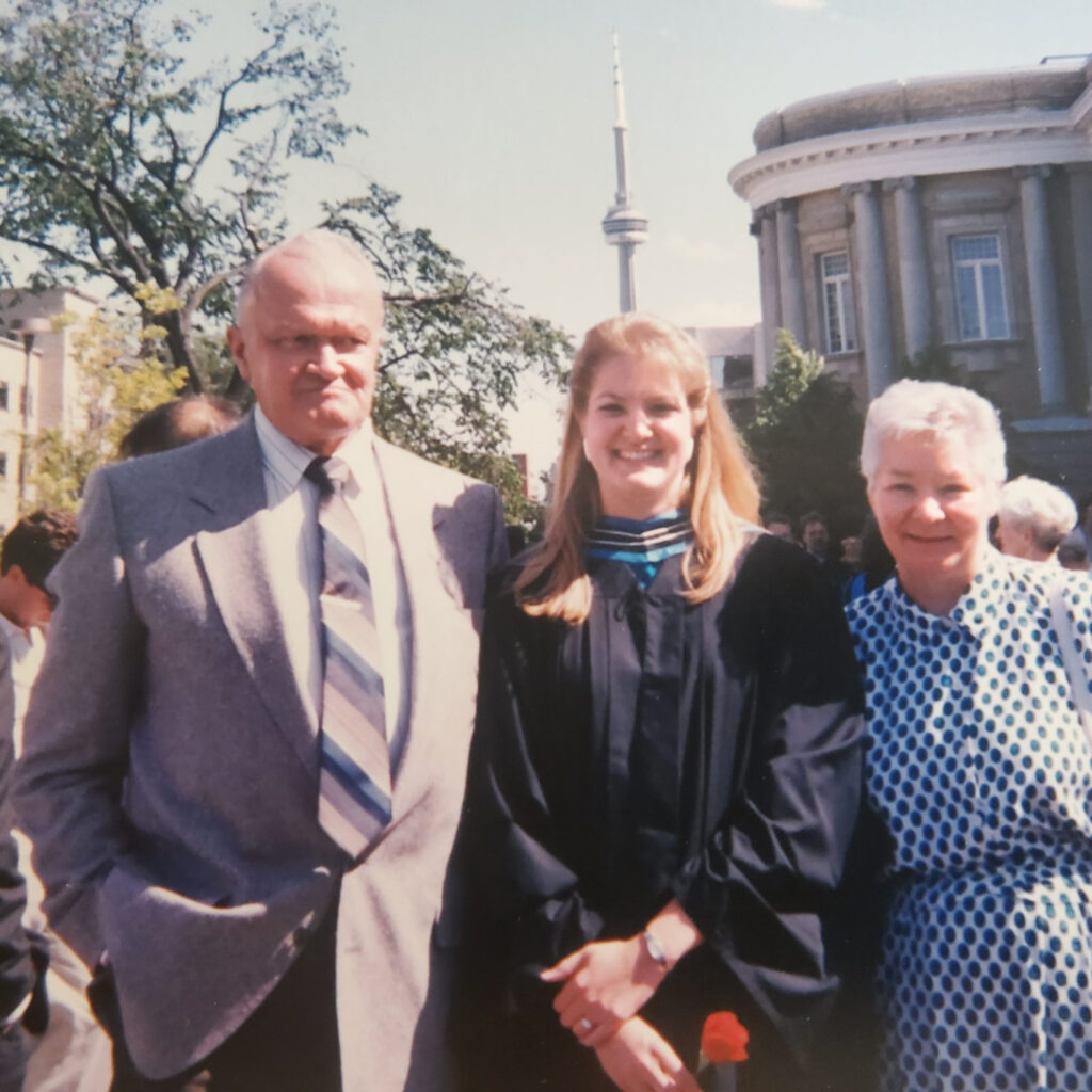 Three people standing in front of Convocation Hall with the CN Tower in the background. Two are parents, one is wearing a graduation gown.