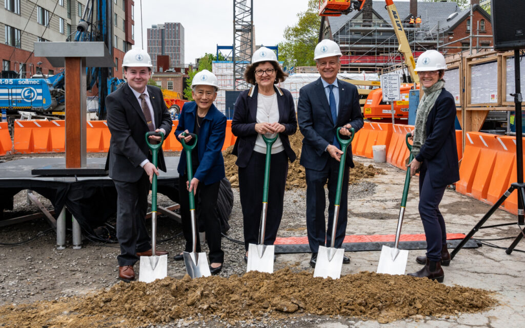 A group of people in business clothing posing for a photo in hard hats and holding shovels at a construction site.