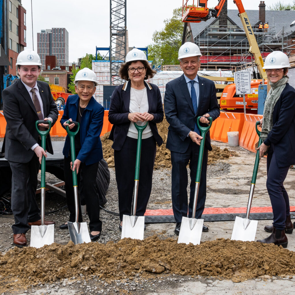 A group of people in business clothing posing for a photo in hard hats and holding shovels at a construction site. 